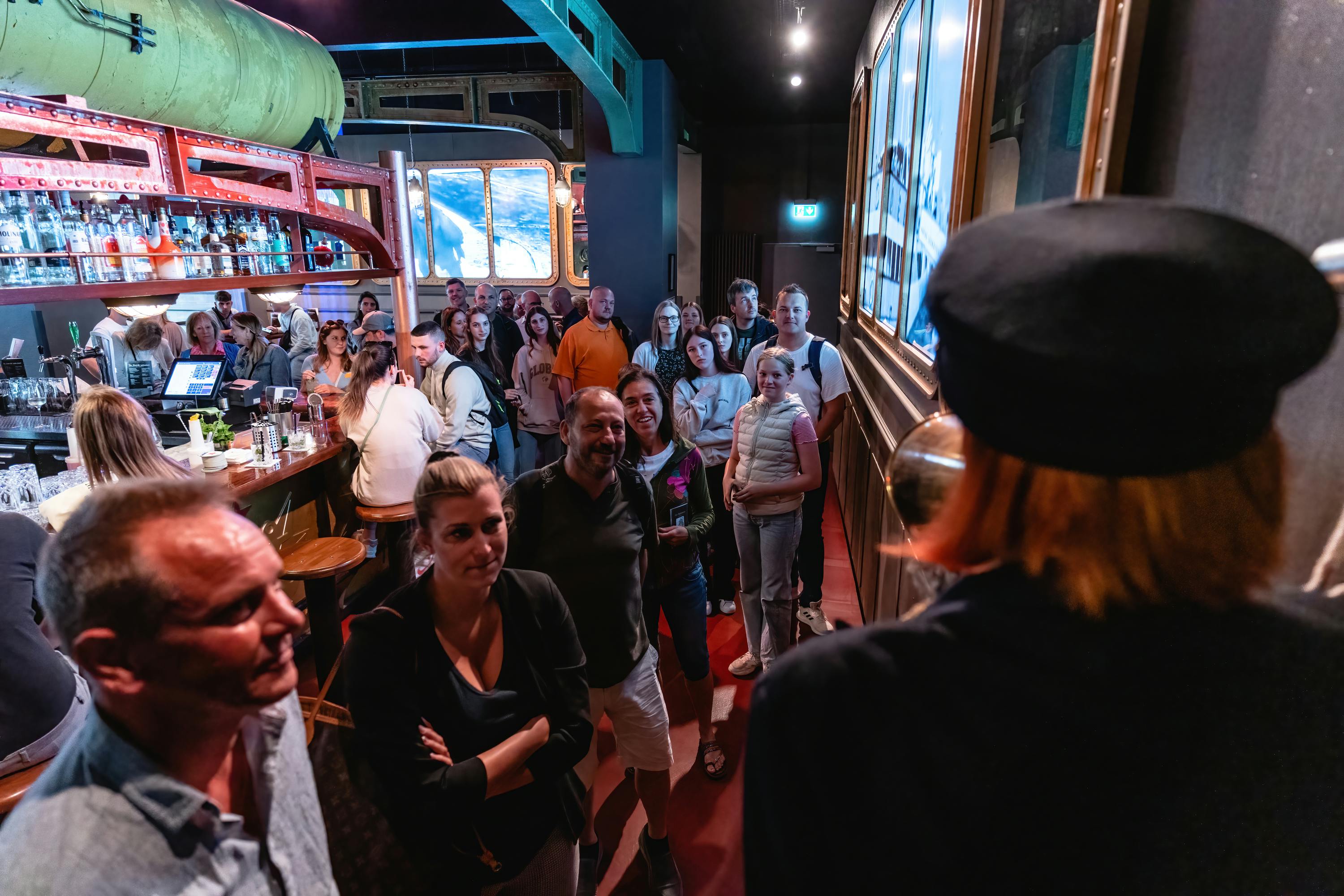 녹색 탱크가 있고, 바 의자, 병이 보이는 바쁜 바에 사람들이 서서 앉아 있습니다. 많은 인파가 몰려 있습니다.