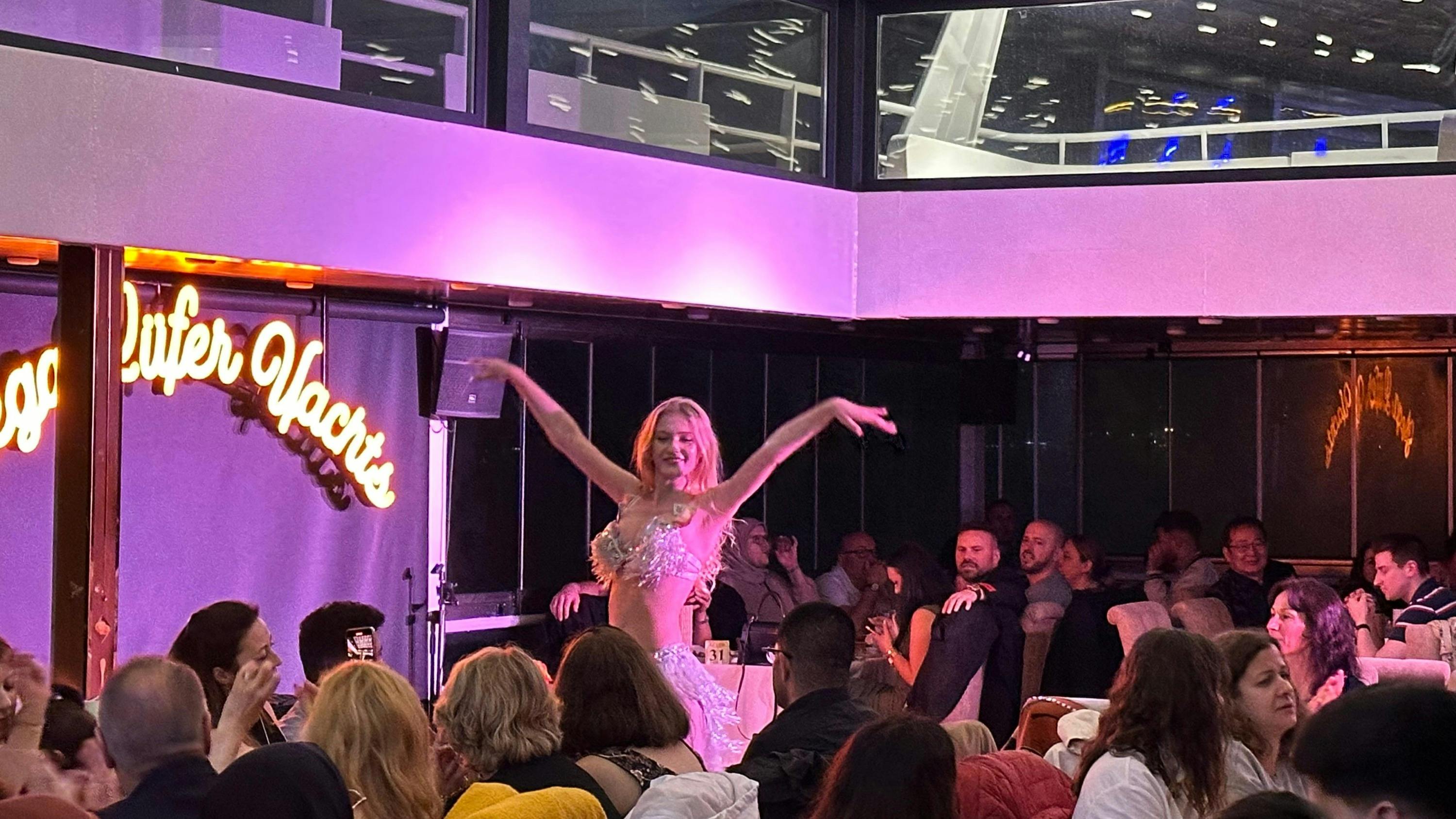 A dancer performs in front of an audience seated at tables in a dimly lit indoor venue with purple lighting.