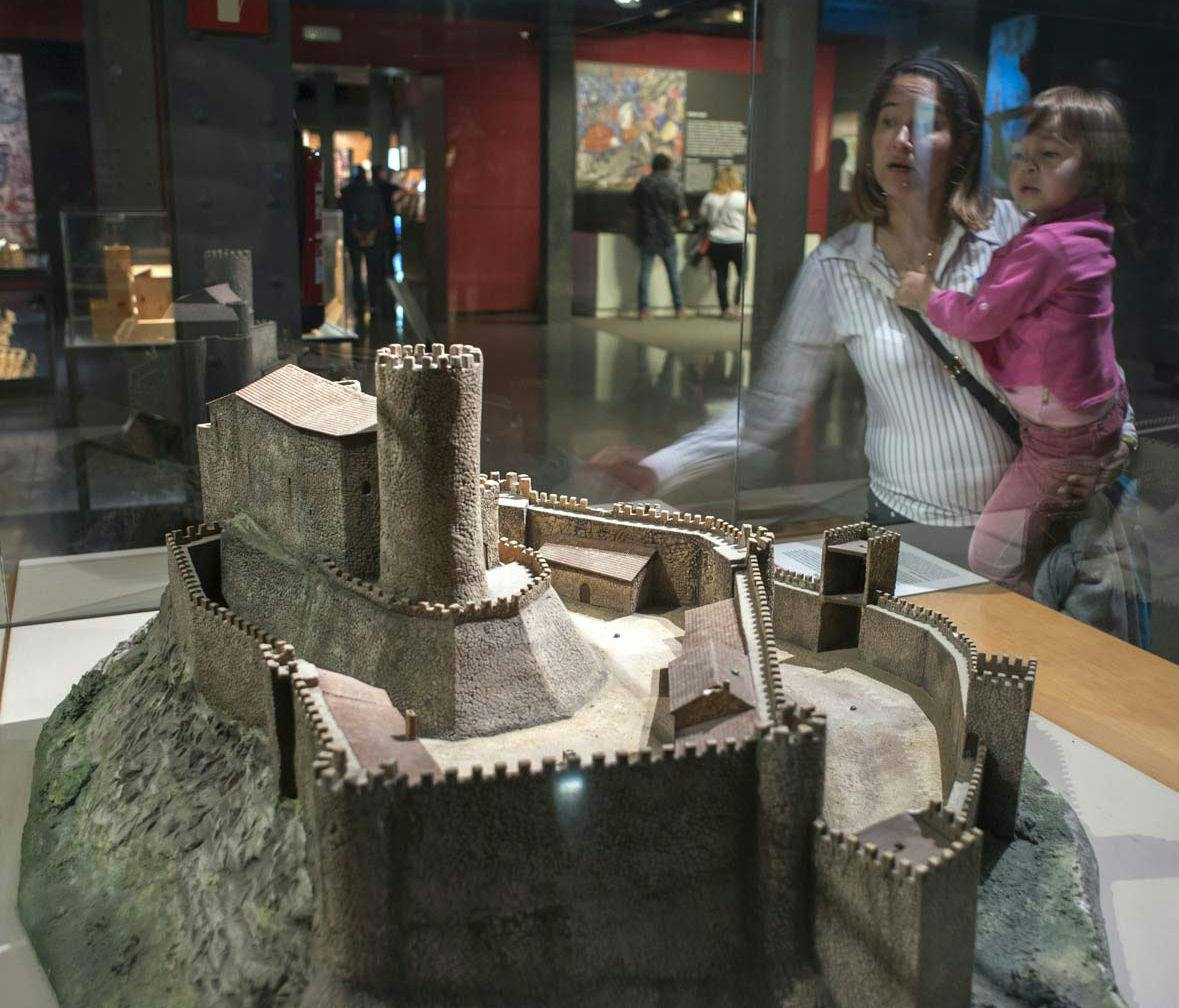 A woman holding a child looks at a detailed miniature model of a medieval castle in a museum setting.