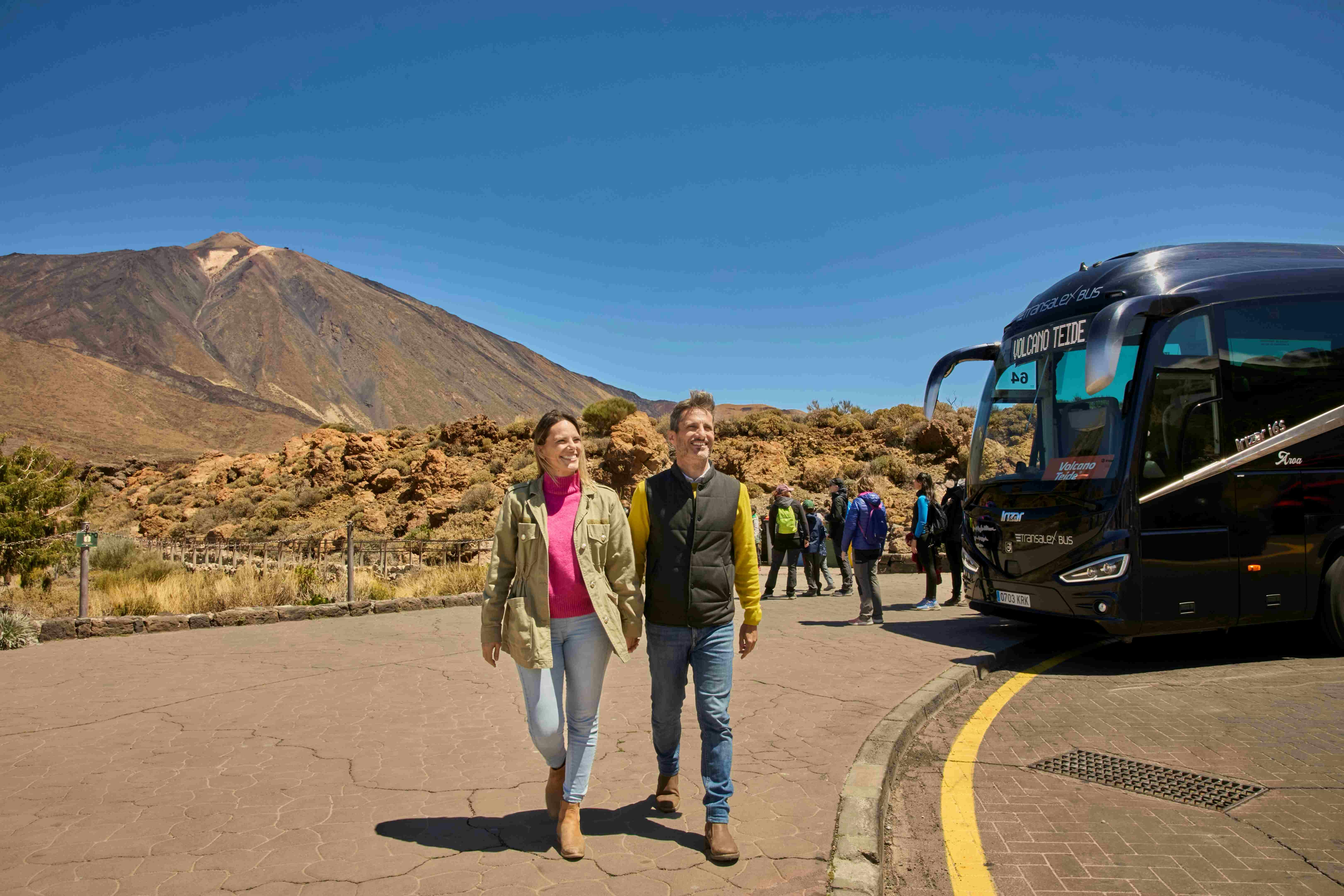 A couple walks on a paved path with a mountain and clear blue sky in the background. Tourists and a bus are in the background.