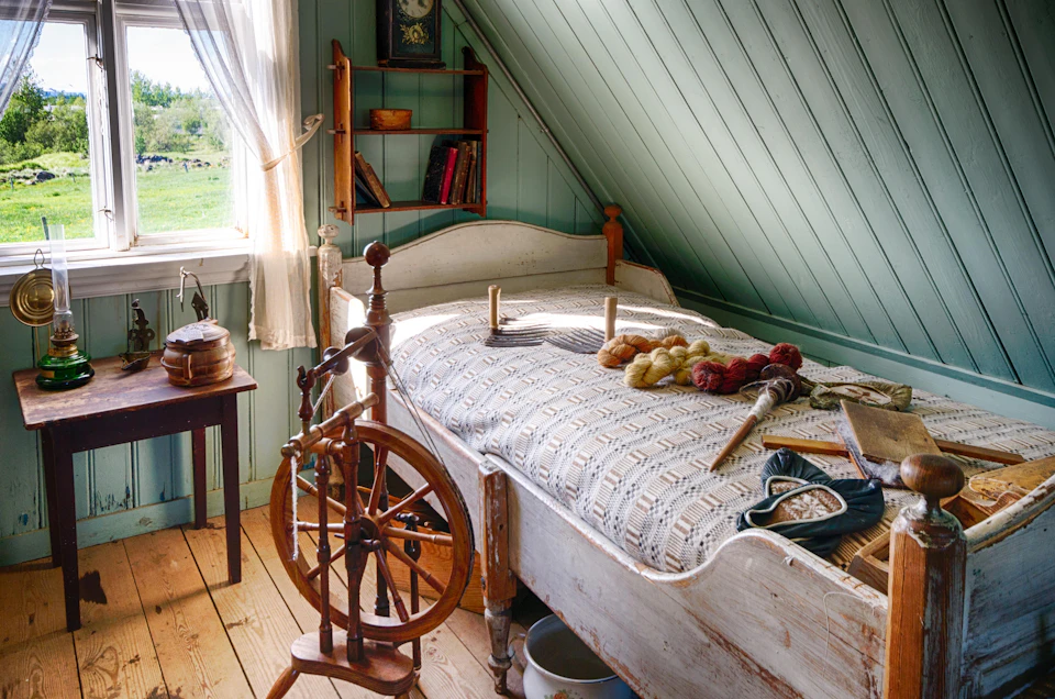 A rustic bedroom with a spinning wheel, a bed covered with a crocheted blanket, yarn, and books on wall shelves by a window.