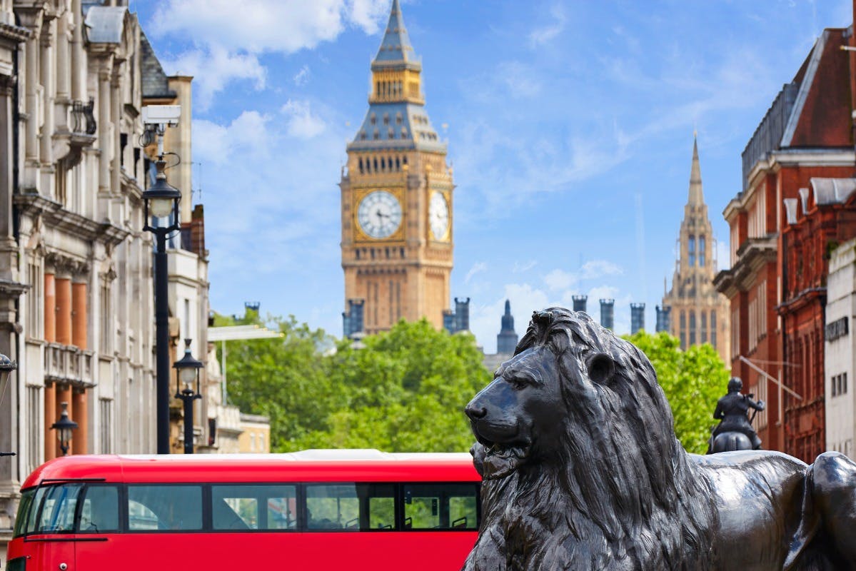 A lion statue in the foreground, a red double-decker bus, Big Ben, and trees under a blue sky in the background.