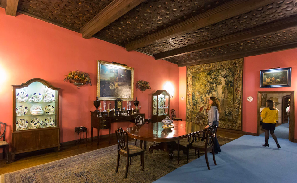 An ornate dining room with a large wooden table, antique furniture, artwork on the walls, and a woman standing near a tapestry.