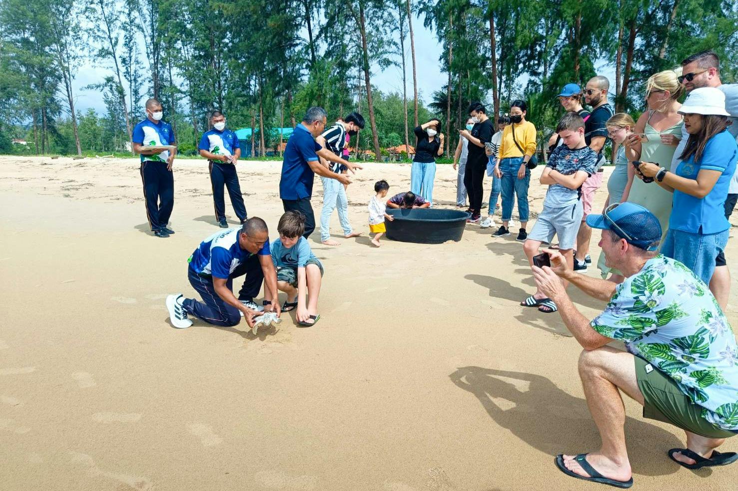 A group of people on a sandy beach, observing and photographing a small child and others near a black container on the ground.