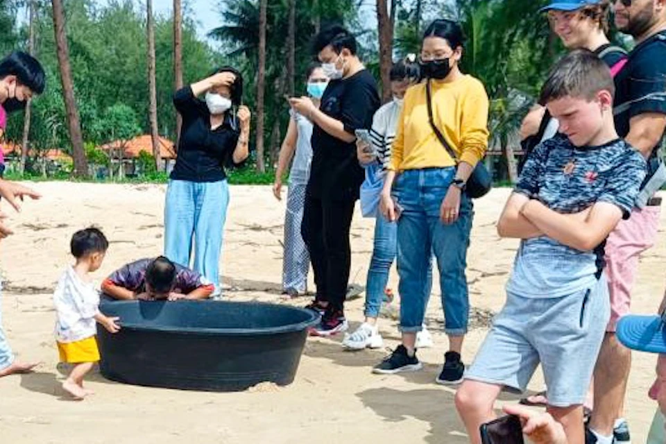 A group of people on a sandy beach, observing and photographing a small child and others near a black container on the ground.