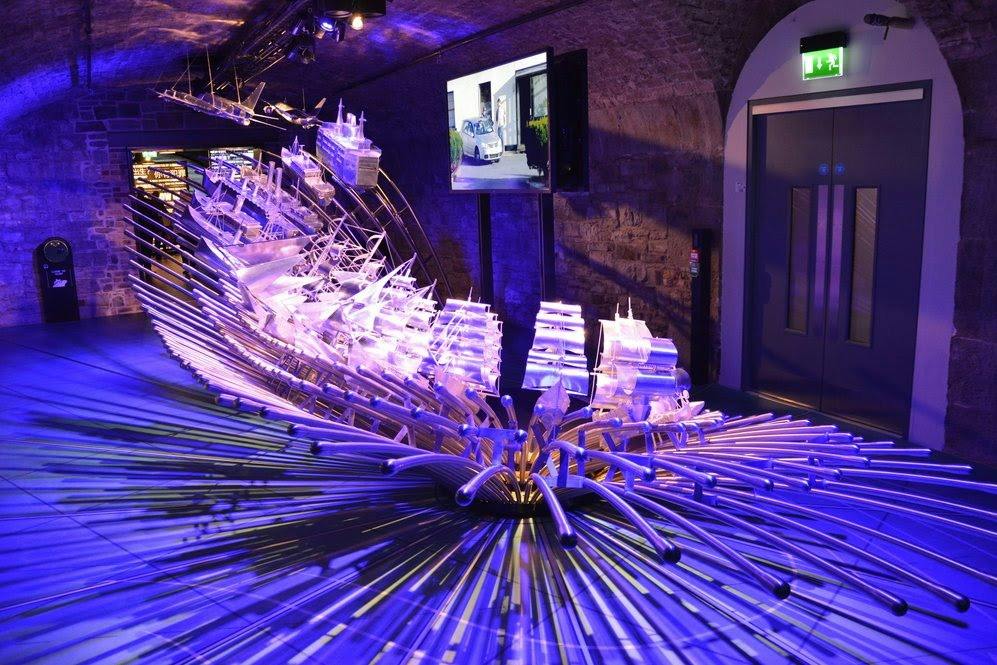 An illuminated art installation featuring stylized ships and rods under purple lighting in a dimly lit indoor space.
