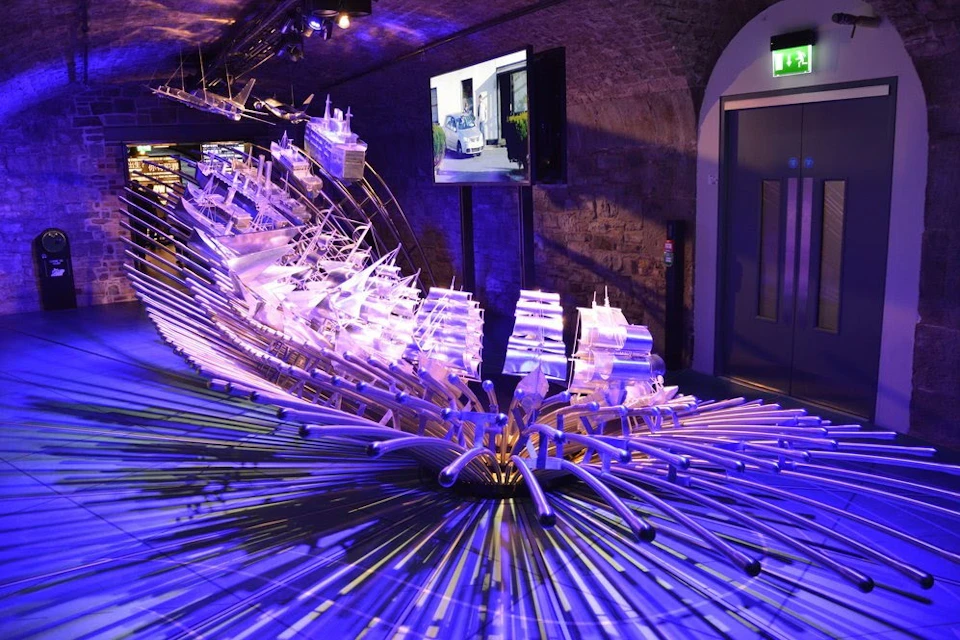 An illuminated art installation featuring stylized ships and rods under purple lighting in a dimly lit indoor space.
