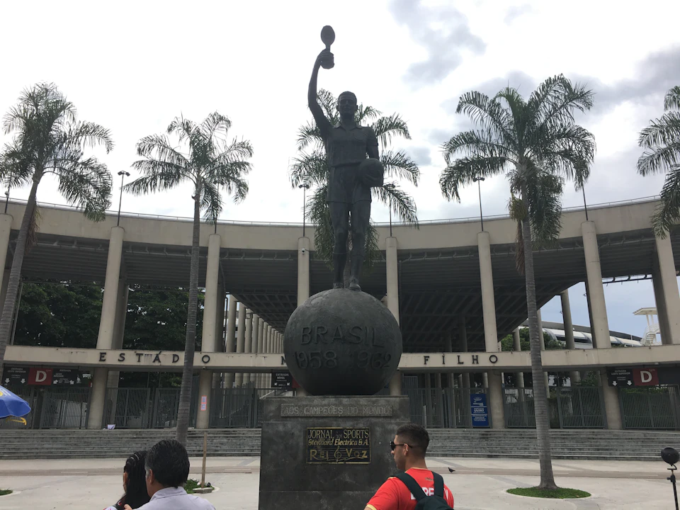 Bronze statue of a soccer player holding a trophy, set against a stadium with palm trees and a few people in the foreground.