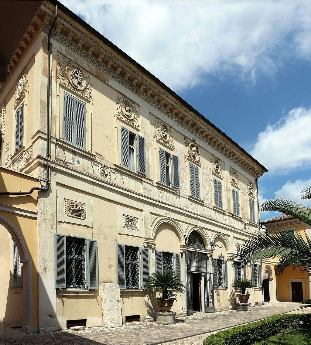 En historisk byggnad med utsmyckade dekorationer, välvd ingång och flera fönster med fönsterluckor. Palmplantor och klar himmel.