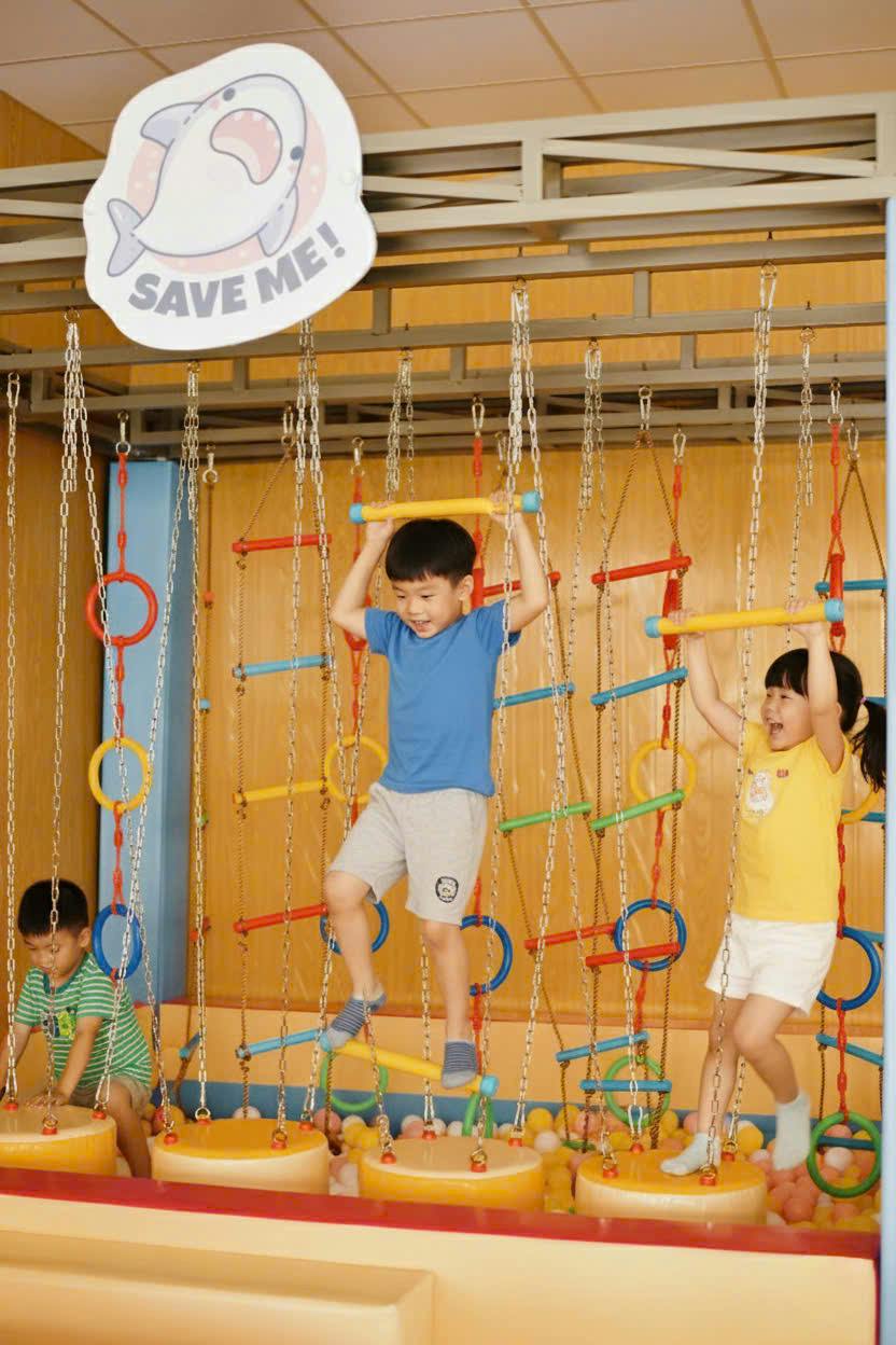 Three children playing on a colorful indoor climbing structure with hanging chains, rings, and bars.