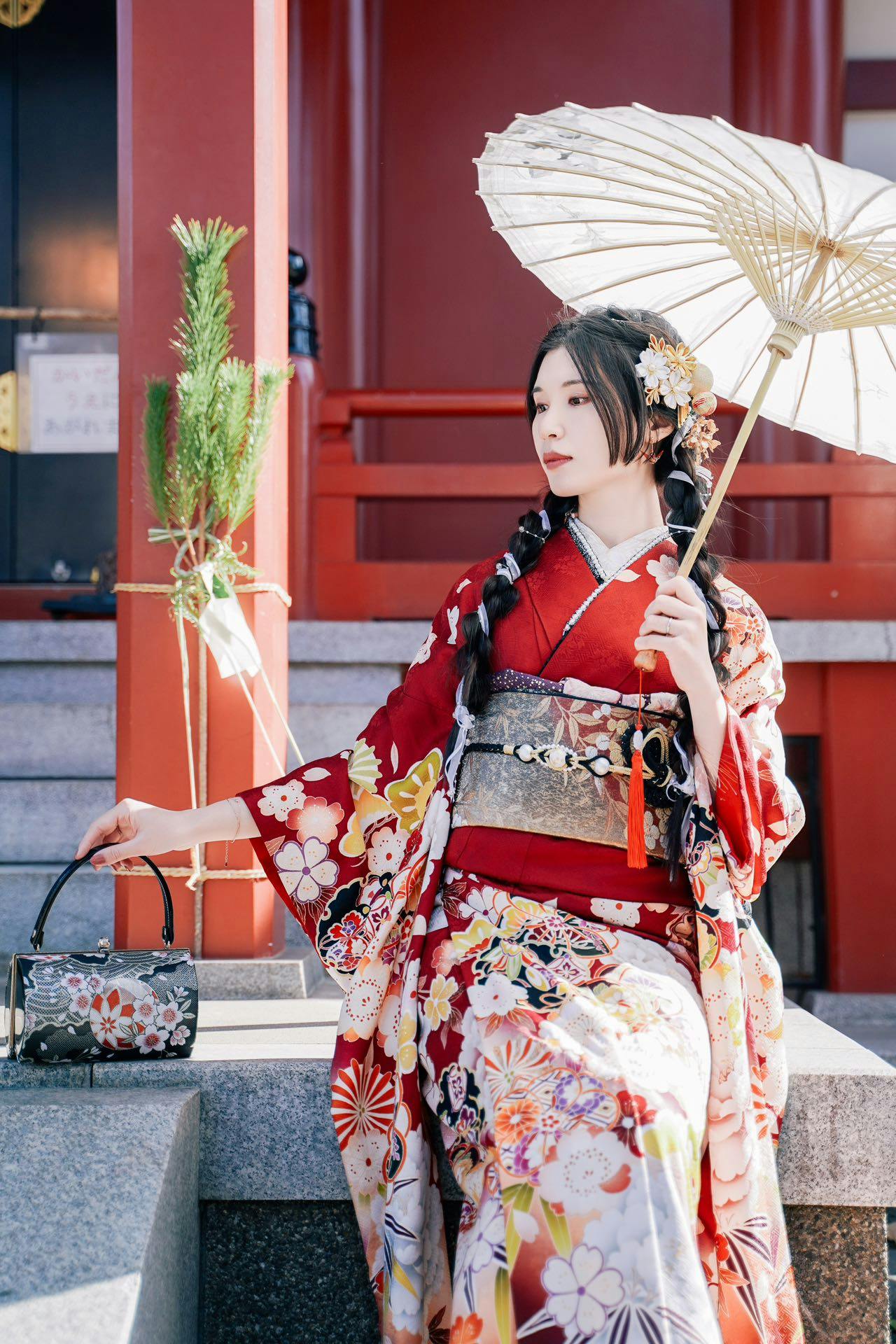 A woman in a red floral kimono holding a white parasol, standing in front of a traditional building with red accents.