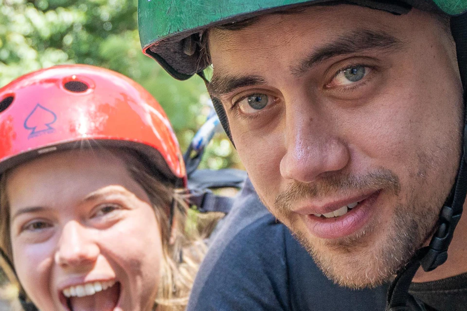 Two people in helmets, harnessed for an outdoor adventure activity, smile at the camera. One gives a peace sign. Trees are in the background.