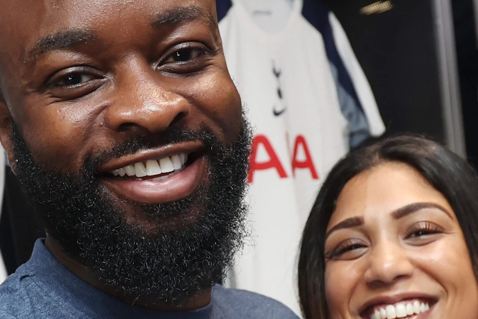 A smiling man and woman take a selfie in a locker room with Tottenham Hotspur jerseys hanging in the background.
