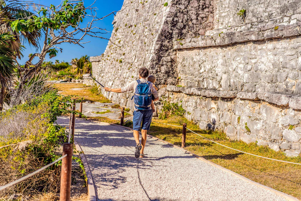 A person carrying a child walks along a path near an ancient stone wall under a clear blue sky.