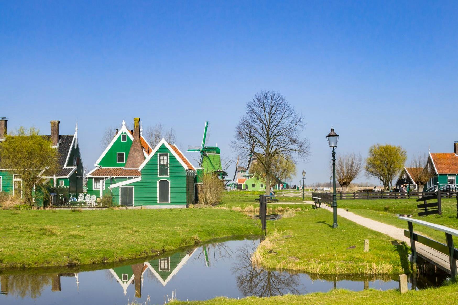 小さな運河の近くにある緑の家と風車小道に沿って木と街灯柱があり、すべて澄んだ青い空の下にあります。