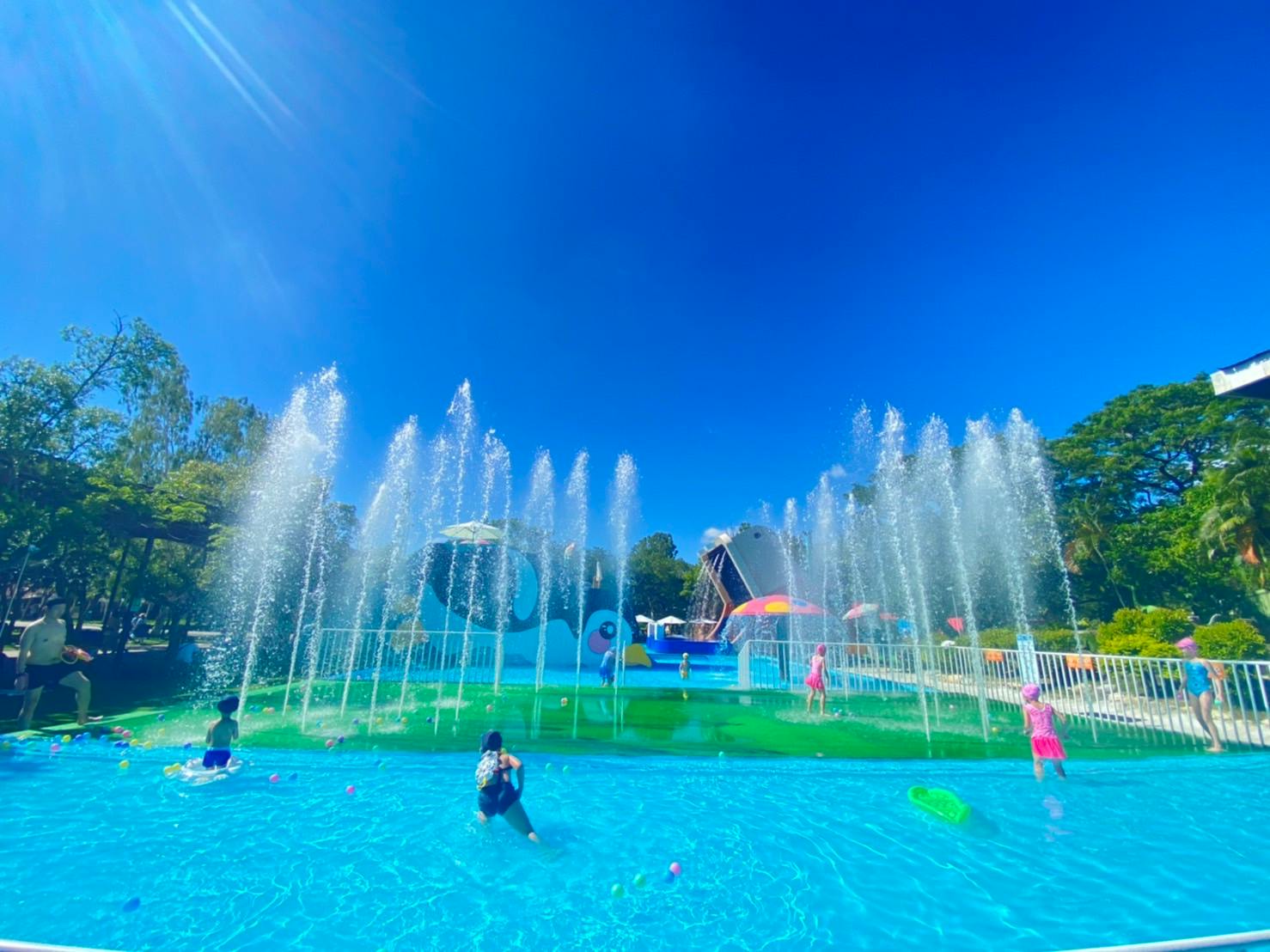 Children playing in a water park with fountains spraying water under a clear blue sky.