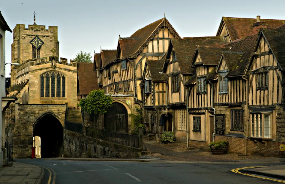 Street with medieval timber-framed buildings alongside a stone archway, with a tree and small plants in front.