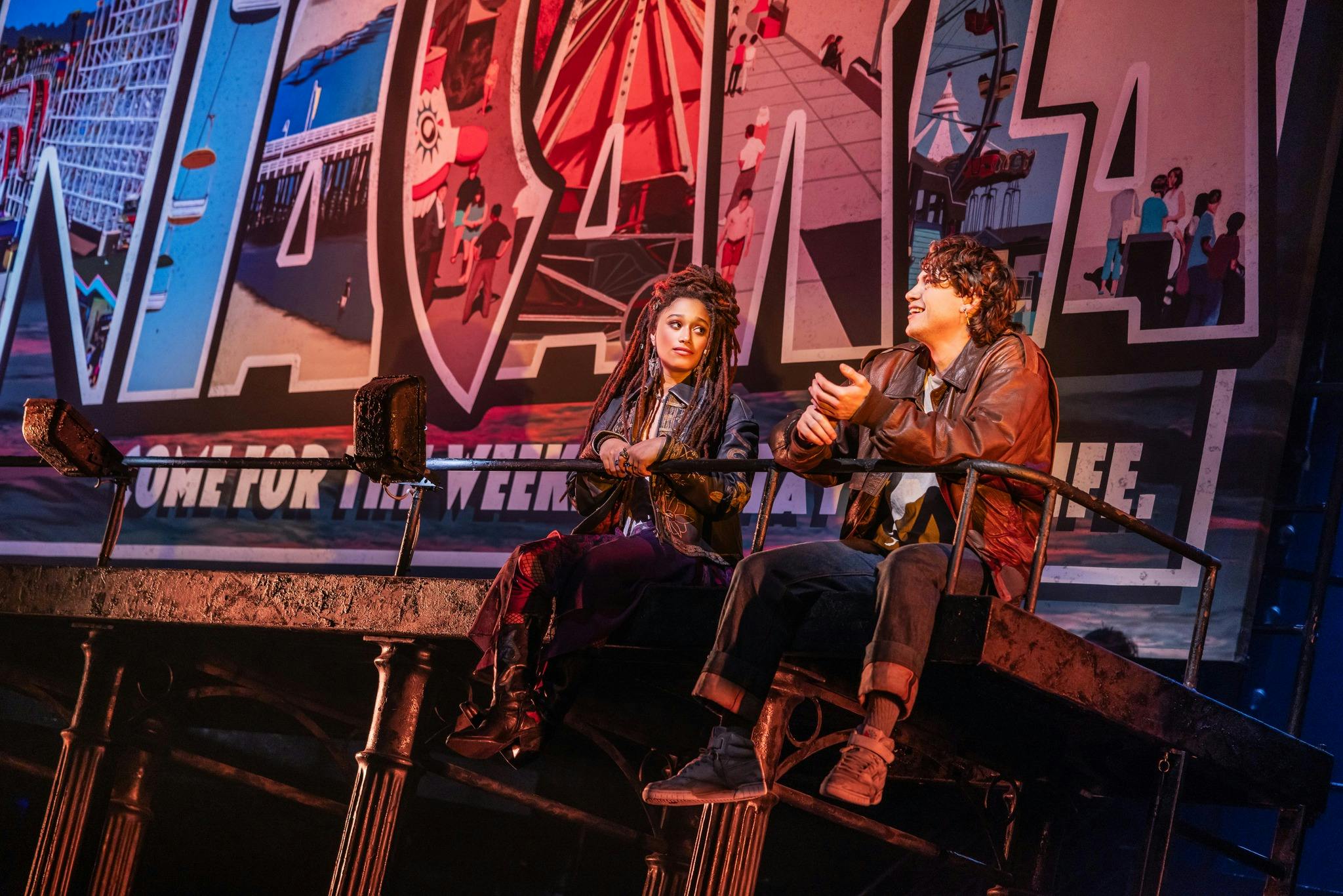 Two people sitting on a raised platform in front of a colorful backdrop with carnival illustrations and large text.