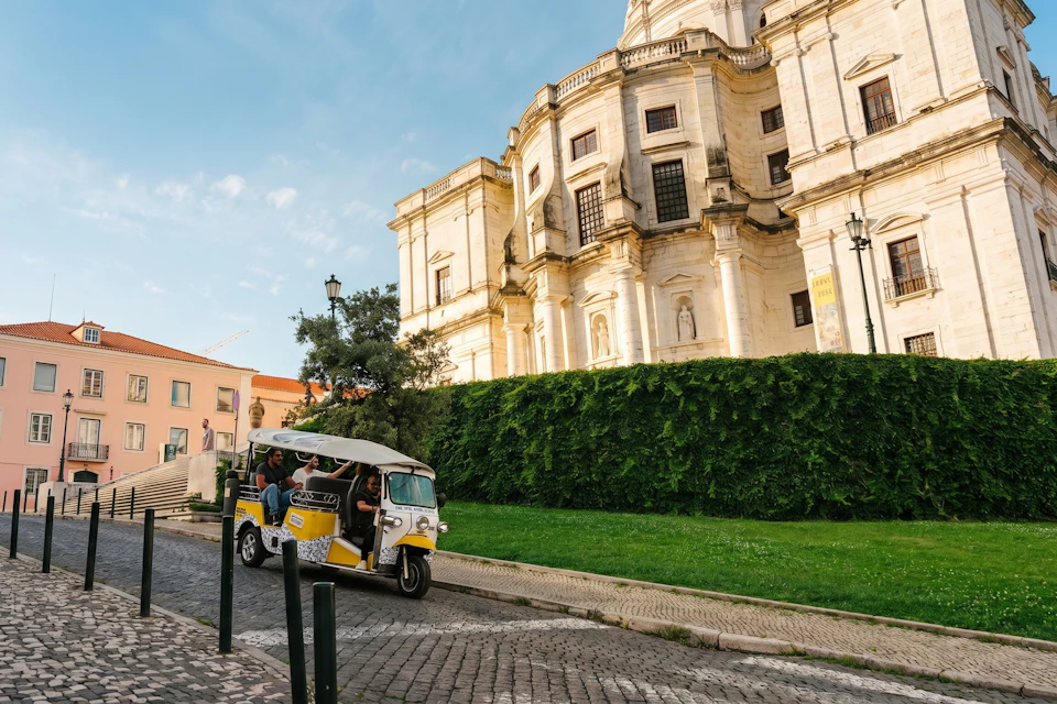 A tuk-tuk with passengers driving on a cobblestone street near a historic building with tall green hedges in the background.