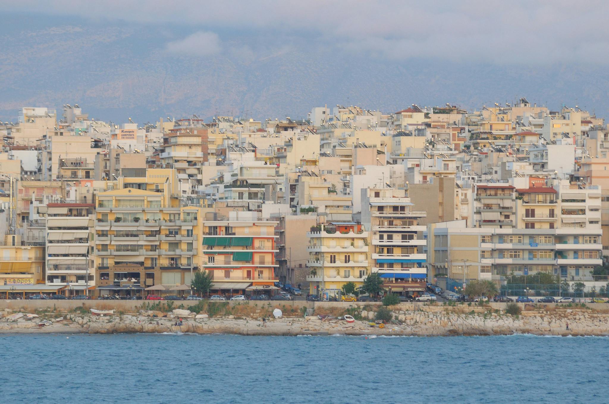Flera mellanhöga flerbostadshus vid vattnet, med berg i bakgrunden och en stenstrand i förgrunden.