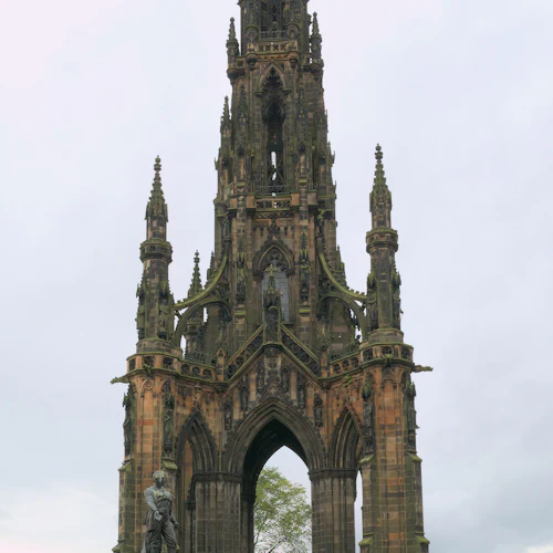 Ett högt, detaljerat gotiskt monument med valv i parkmiljö, med en staty vid basen och en molnig himmel ovanför.