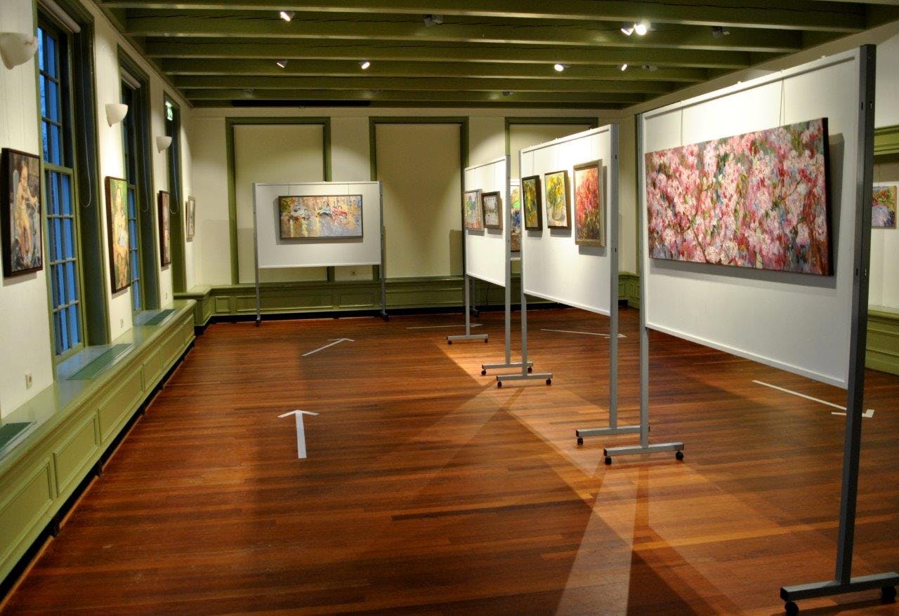 흰색 칸막이 벽에 나무 바닥, 녹색 천장 대들보, 다양한 액자 그림이 있는 미술관. 바닥에 화살표 표시.