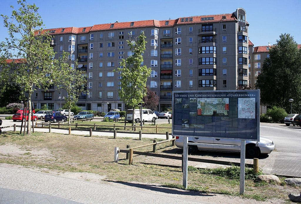 A parking lot with cars, trees, and an informational sign in front of a multi-story residential building with a red roof.