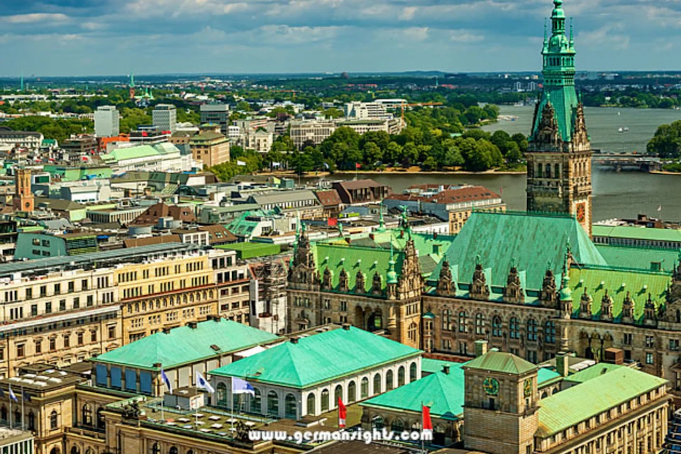 Aerial view of a cityscape with historic buildings, green rooftops, a tall tower, and a river in the background under a partly cloudy sky.