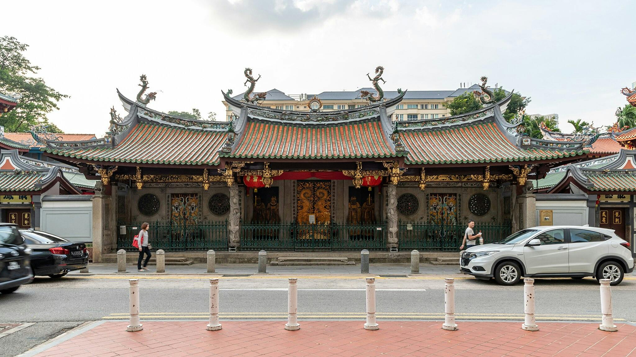 A traditional temple with decorative roof carvings, red lanterns, and intricate doors, flanked by parked cars and pedestrians.