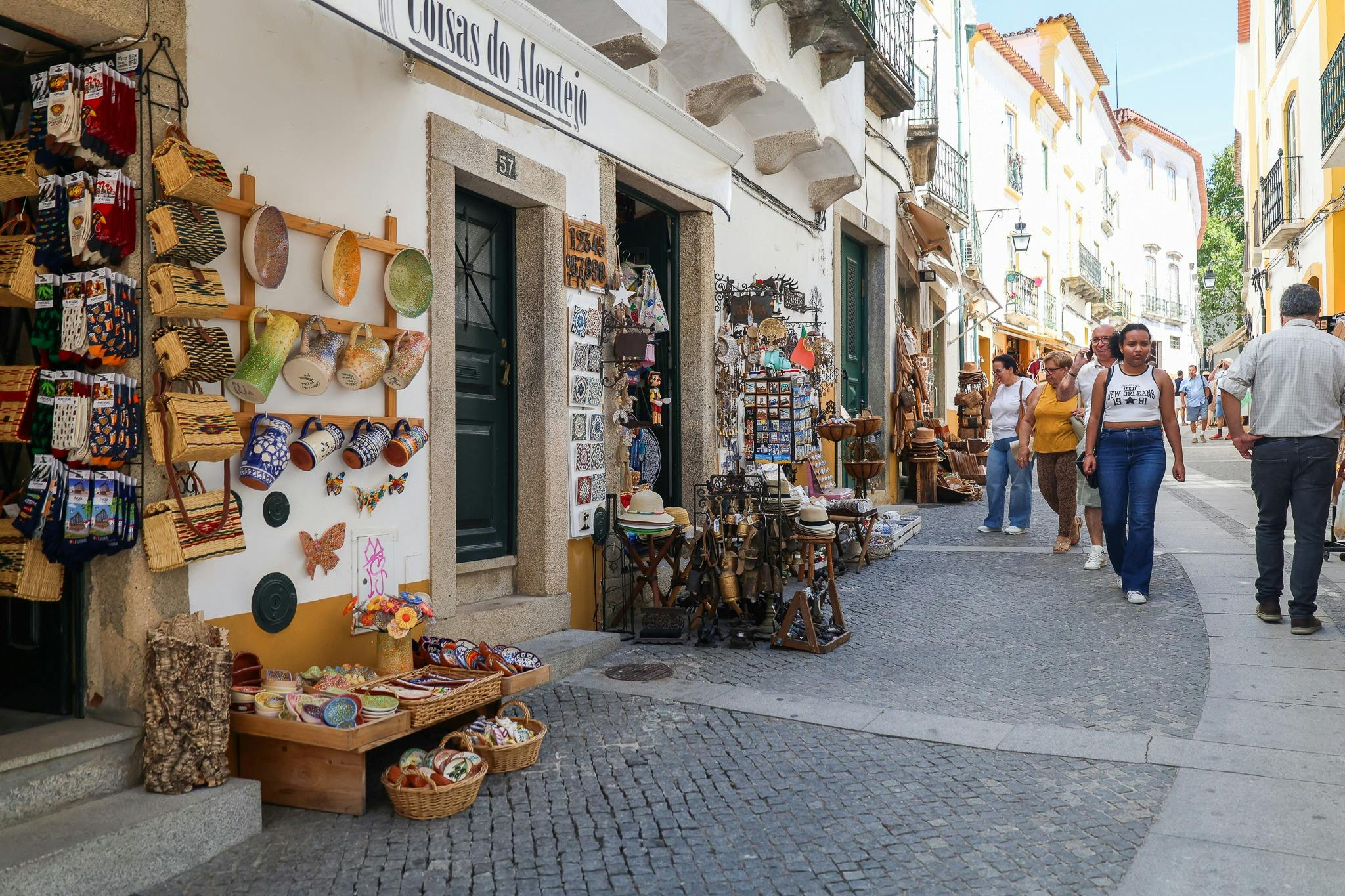 People walk on a narrow, cobblestone street lined with shops displaying various goods and souvenirs in a European town.