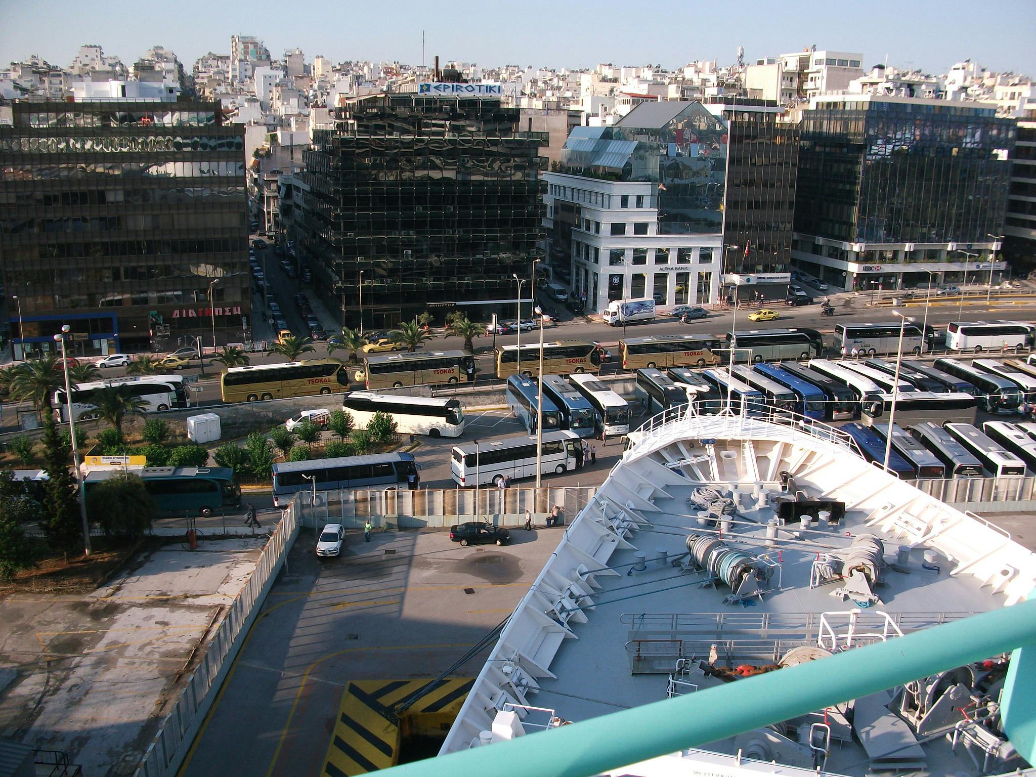 En livlig stadsmiljö med många bussar uppradade, i anslutning till ett fartygsdäck och omgiven av höga byggnader.