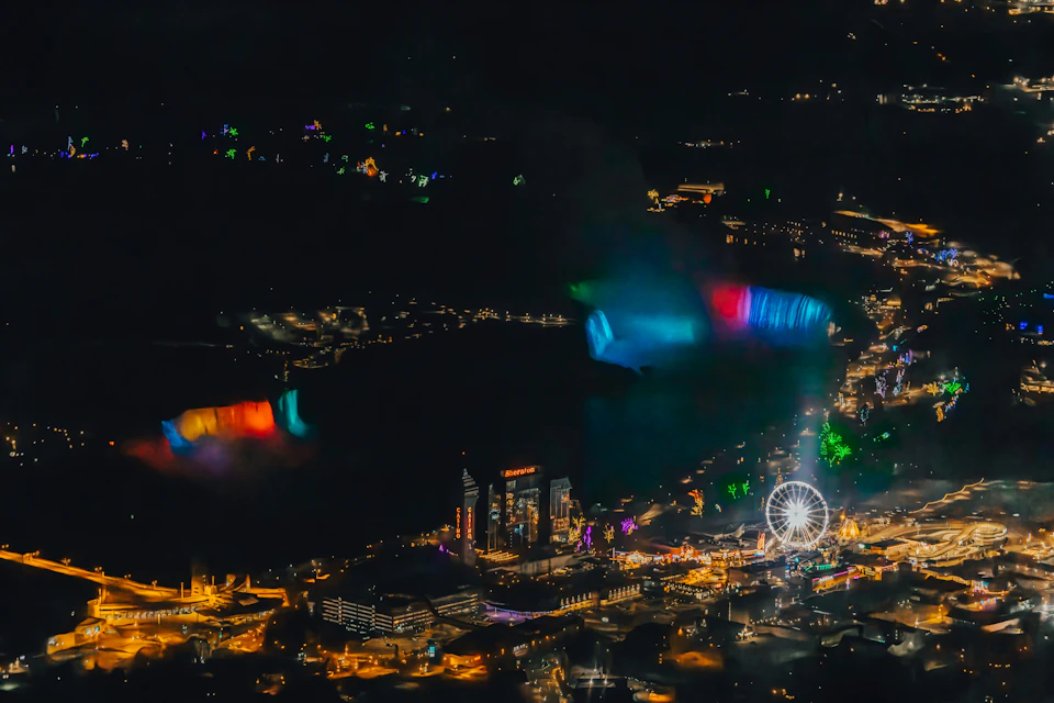 Aerial view of a brightly lit fair with a Ferris wheel, colorful light displays, and surrounding buildings at night.
