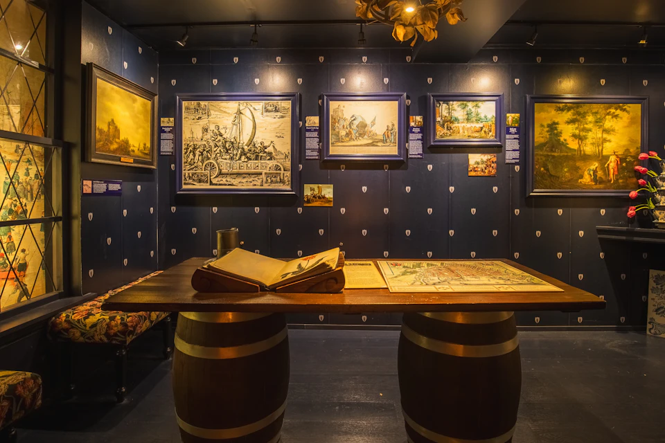A museum display featuring paintings on dark walls and a table with open books on wooden barrels as legs.