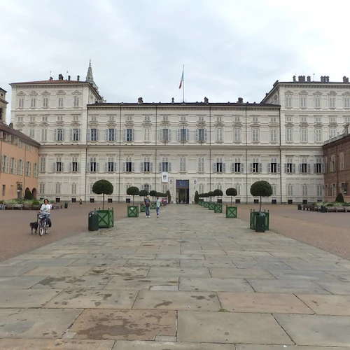 Stor, utsmyckad byggnad med symmetrisk arkitektur, flagga på toppen, bred stenlagd innergård, människor som går och cyklister till vänster.