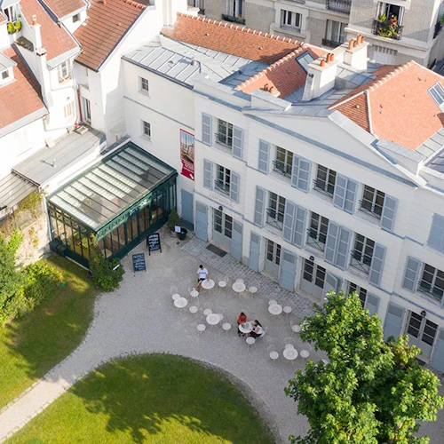 Flygfoto över en innergård med flera bord och stolar, omgiven av vita byggnader med röda tak och en inglasad yta.