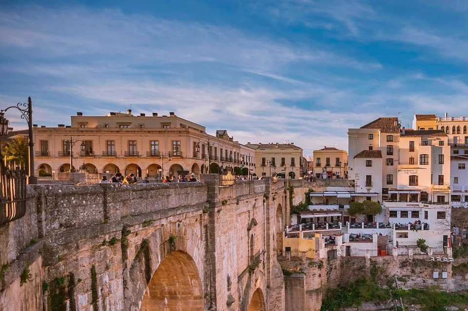 A scenic view of a stone bridge with arches, historic buildings, and people gathered along the bridge under a blue sky.