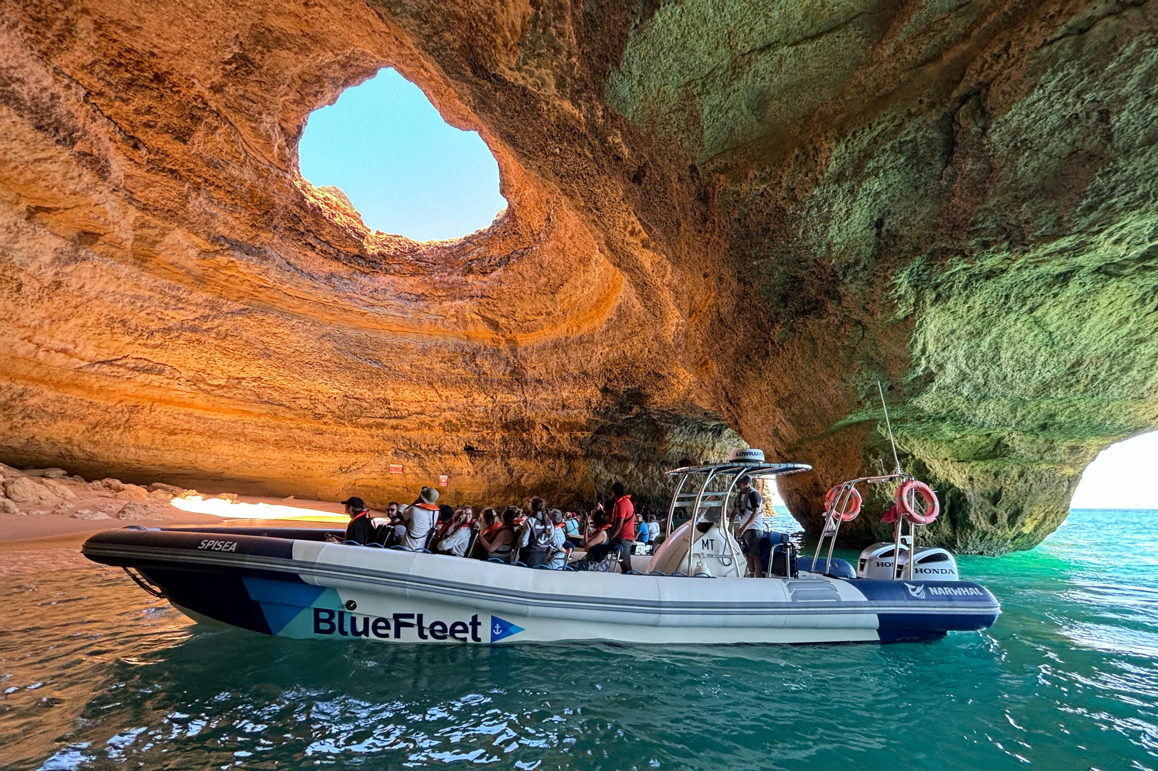 大きな円形の開口部から太陽の光が差し込む岩窟の中、ボートに乗った人々のグループ。