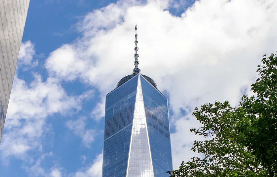The top of the One World Trade Center building against a partly cloudy sky, with some green tree leaves in the lower right corner.