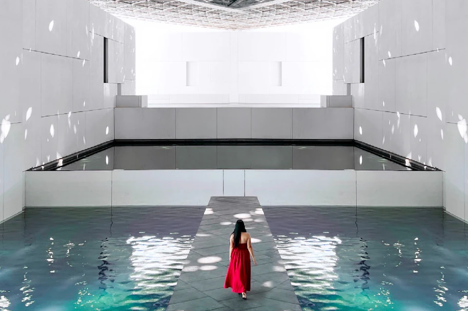 A woman in a red dress walks on a narrow path surrounded by water, inside a modern, white architectural space.