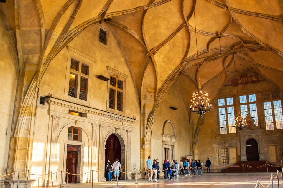 A historic hall with ornate vaulted ceilings, chandeliers, and people walking through, lit by sunlight from large windows.