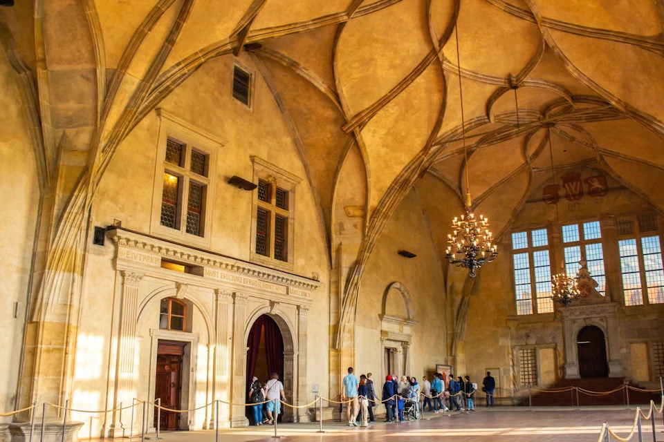 A historic hall with ornate vaulted ceilings, chandeliers, and people walking through, lit by sunlight from large windows.