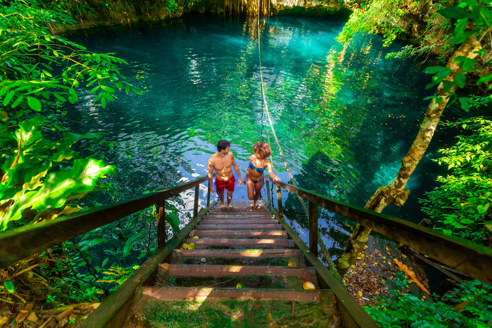 Two people in swimwear ascend wooden stairs from a turquoise water pool surrounded by lush vegetation and sunlight.