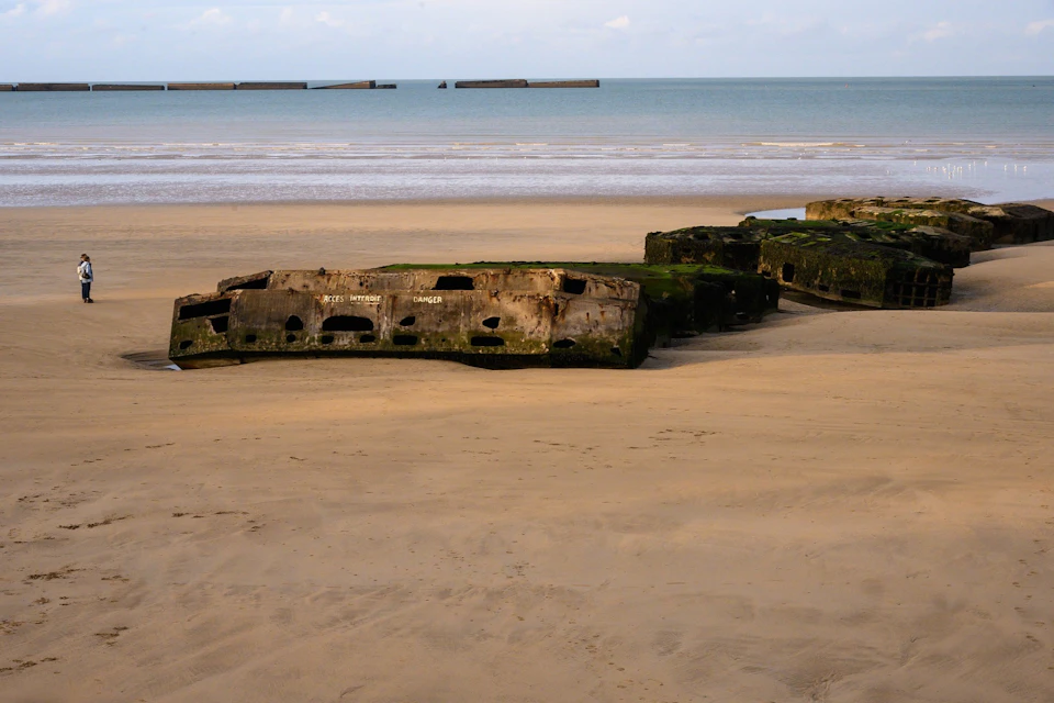Concrete remains of a WWII landing craft on a sandy beach with the sea and sky in the background.