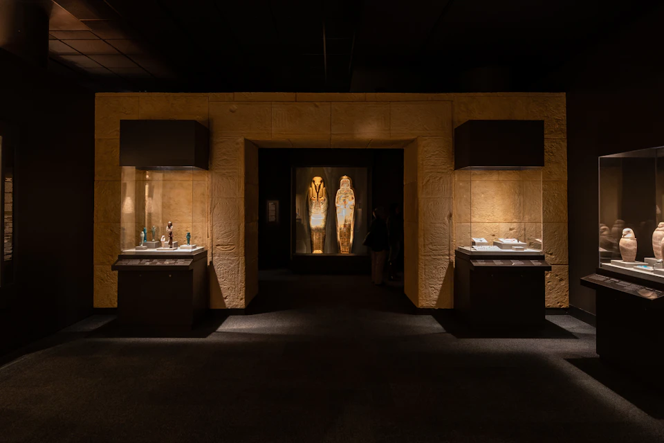 A dimly lit museum exhibit featuring two illuminated Egyptian sarcophagi, with display cases of artifacts on either side.