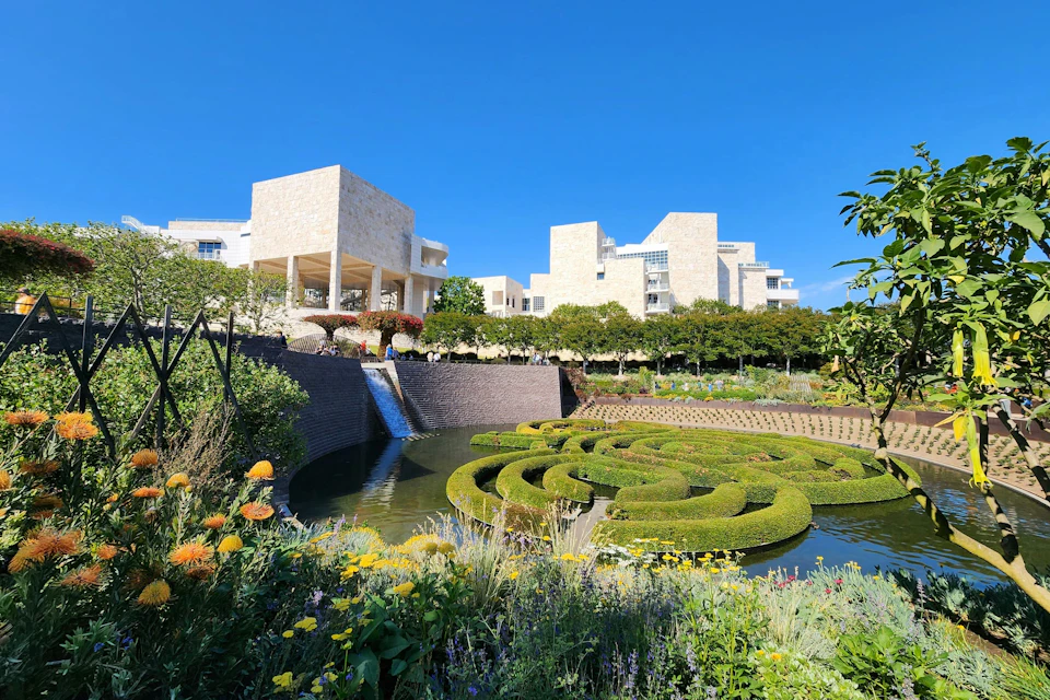 Stone buildings with terraces overlook a manicured garden with a water feature and maze-like hedges under a clear blue sky.