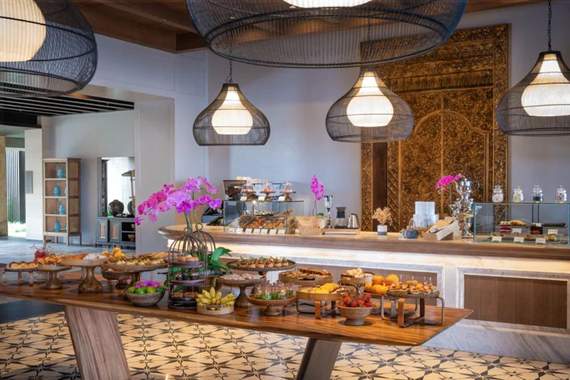 A buffet setup with wooden tables displaying fruits and desserts, decorated with flowers, under hanging lamps in a cozy room.