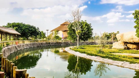 A tranquil pond surrounded by greenery, large rocks, wooden fencing, and a thatched-roof building under a partly cloudy sky.
