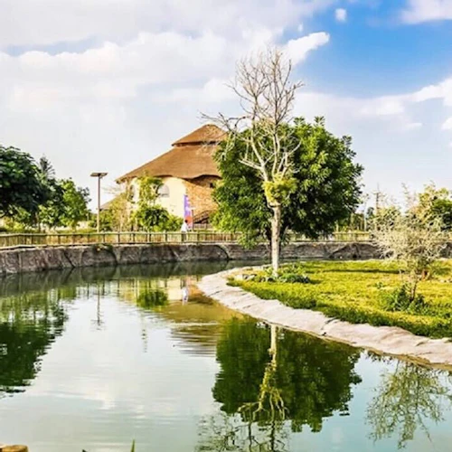 A tranquil pond surrounded by greenery, large rocks, wooden fencing, and a thatched-roof building under a partly cloudy sky.