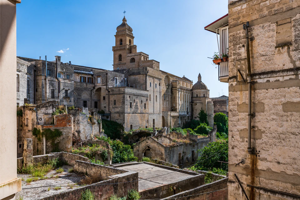 Ancient stone buildings with arches and a bell tower under a clear blue sky, surrounded by overgrown vegetation.