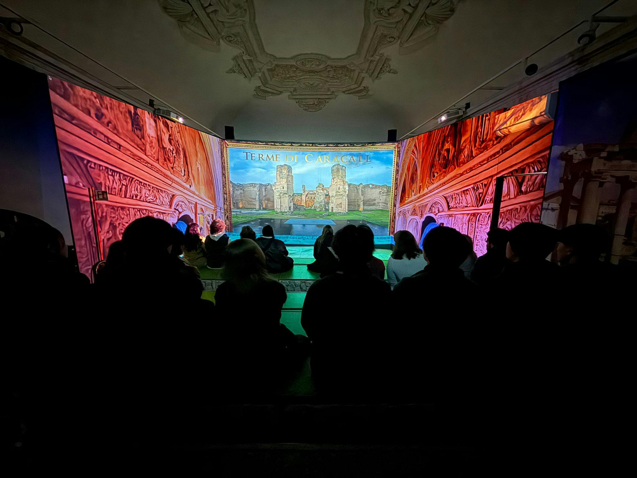 Un gruppo di persone sedute in una stanza buia guarda la proiezione di una scena storica con architettura e testo.